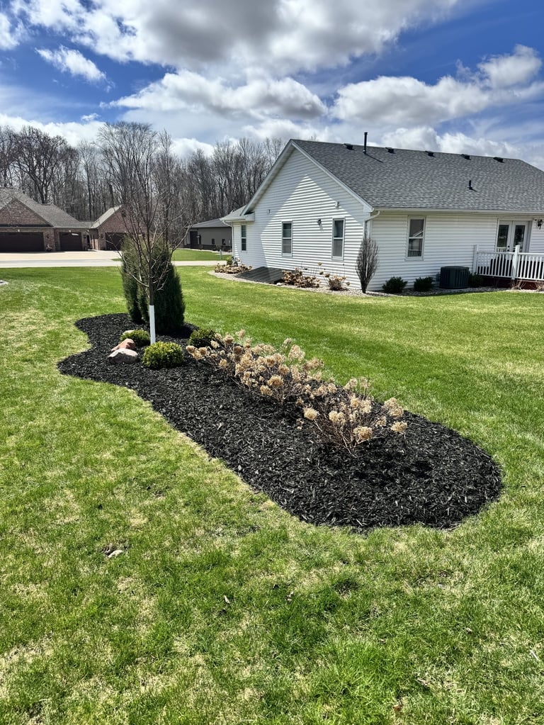 White ranch house with landscaped front yard, mulch flower bed with shrubs, manicured lawn, and bare trees under blue sky