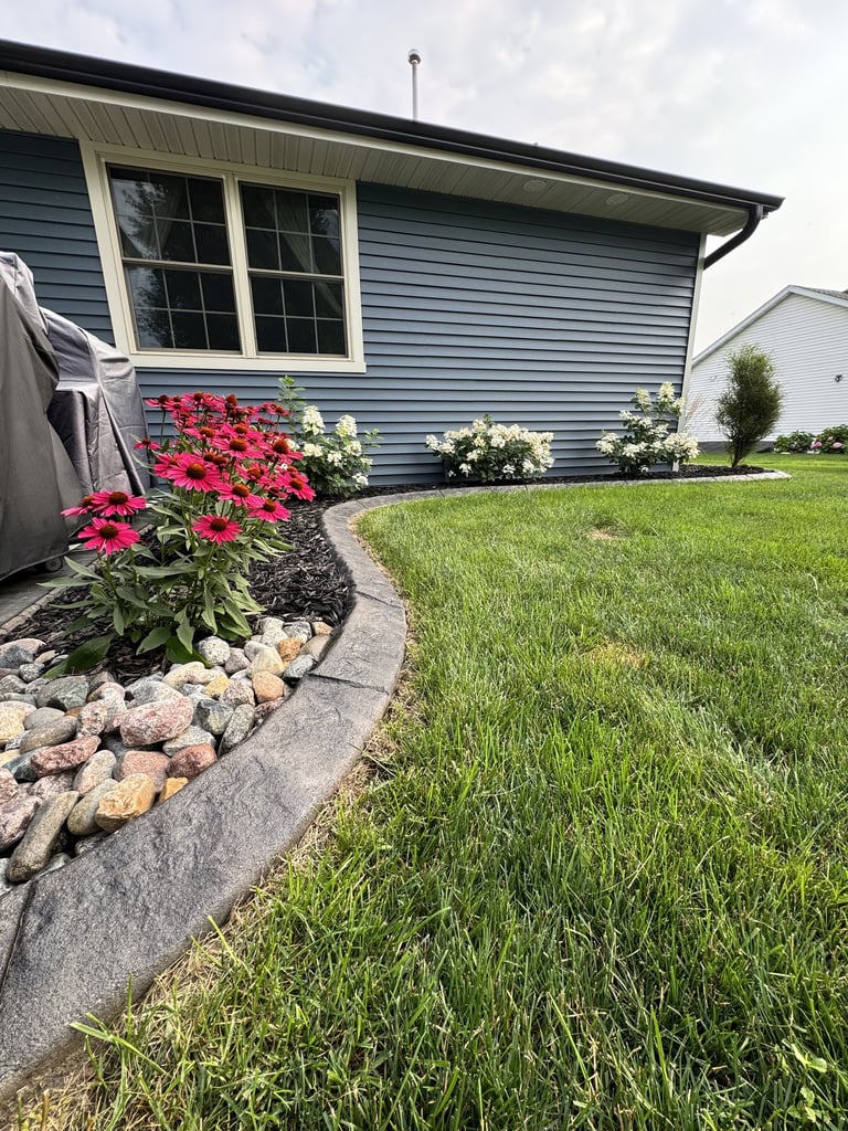 Blue house exterior with red and white flowers in landscaped garden bed edged with stones and metal trim, green lawn