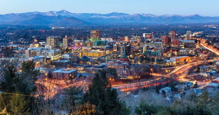 Asheville city skyline at dusk with illuminated buildings, river reflecting lights, snow-capped mountains in background, and evergreen trees in foreground.