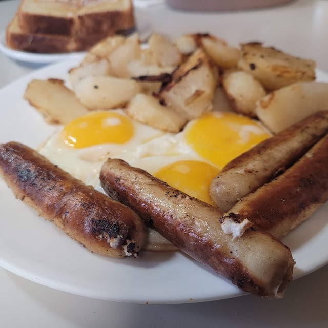 A breakfast plate with scrambled eggs, sausage links, and roasted potatoes, alongside toast in the background