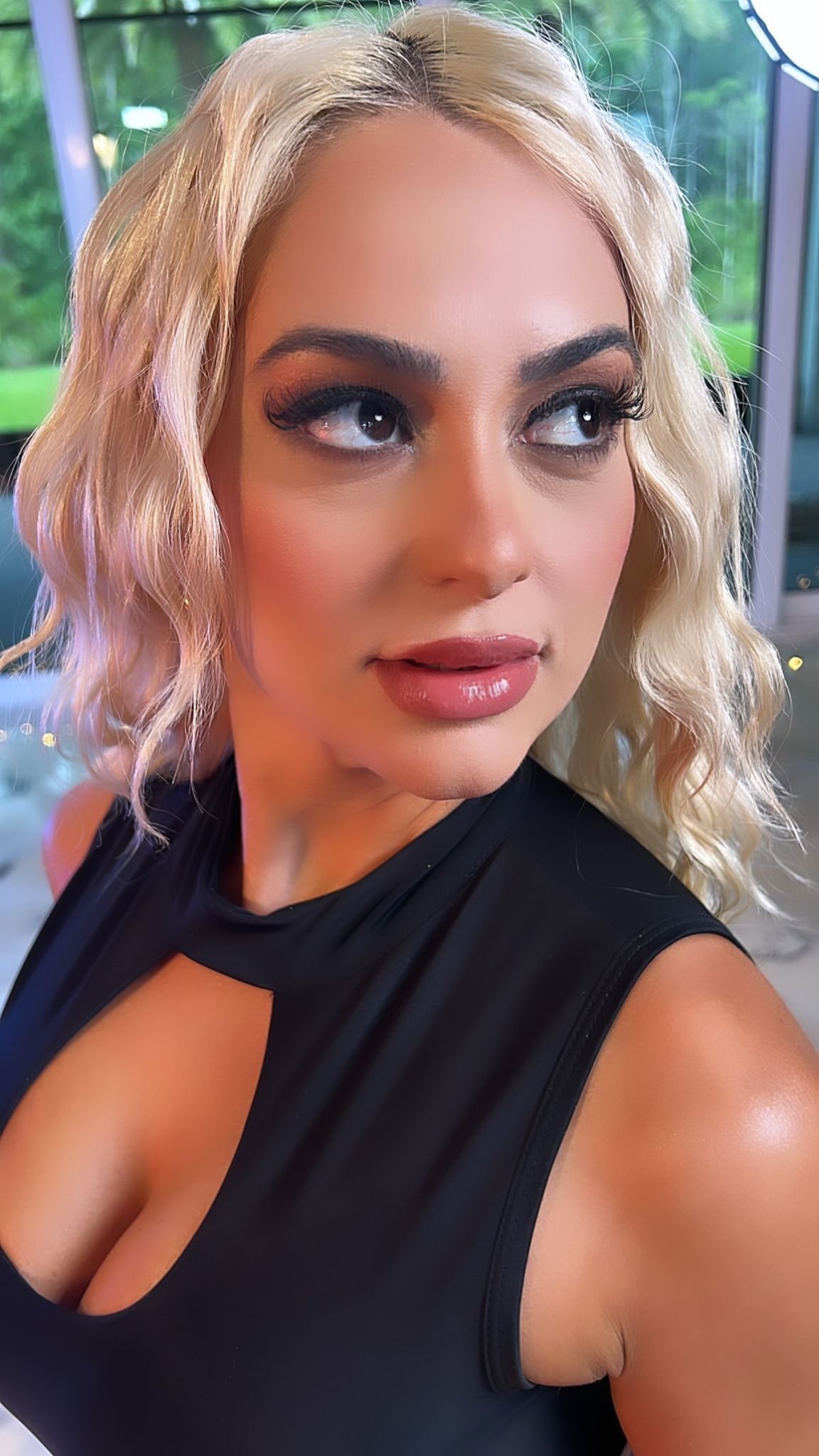 Woman with platinum blonde wavy hair and bold makeup wearing a black sleeveless top, looking at camera