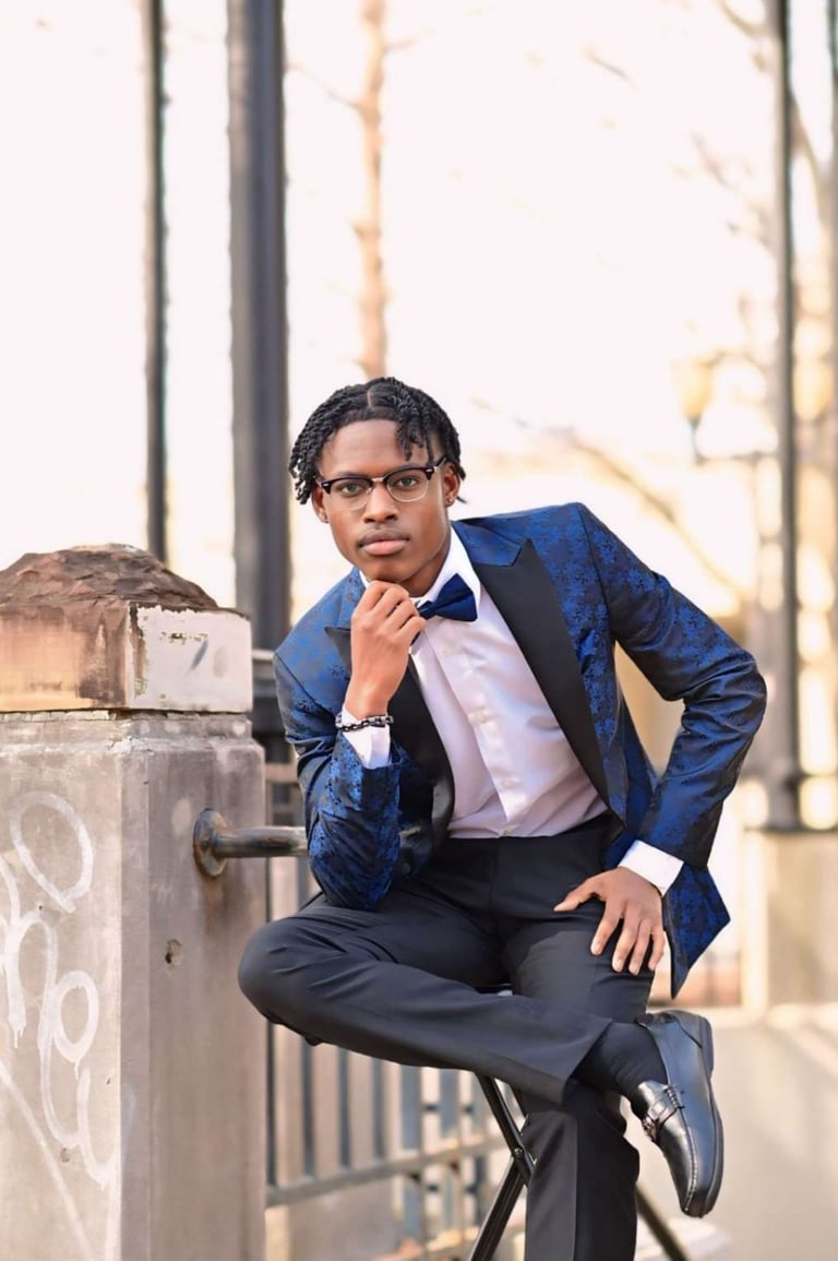 Young man in blue blazer and glasses sitting on concrete structure outdoors, posing thoughtfully