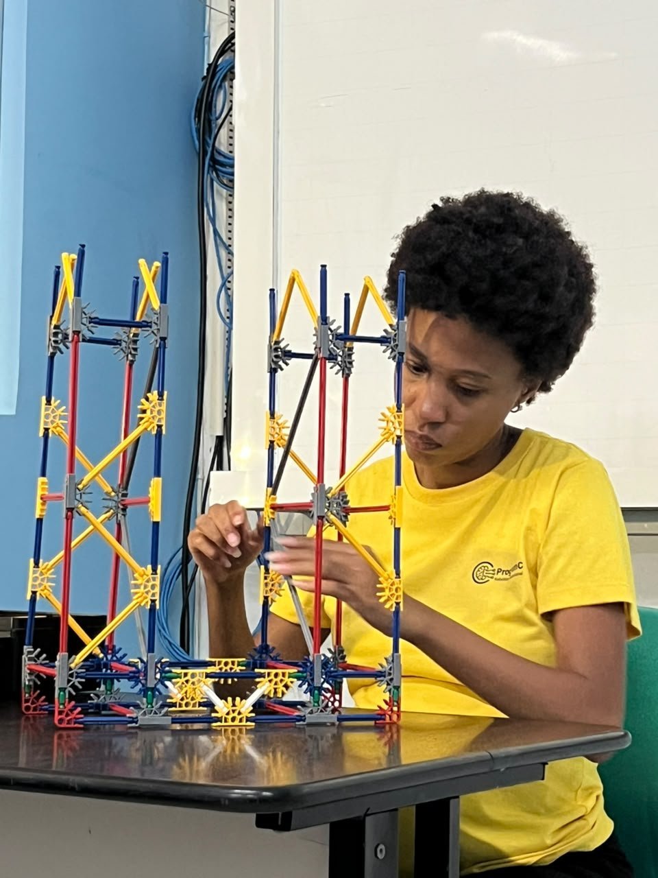 Aluna da ProgramC em camisa amarela montando estrutura geométrica com hastes e conectores
