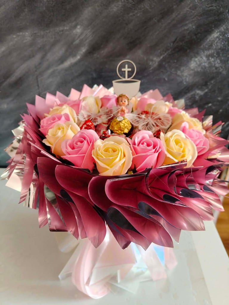 Kompozycja pierwszokomunijna z różowymi i kremowymi różami, złotą Ferrero Rocher, aniołkiem i złotym krzyżykiem na szczycie, w białym wazonie z bordowymi płatkami