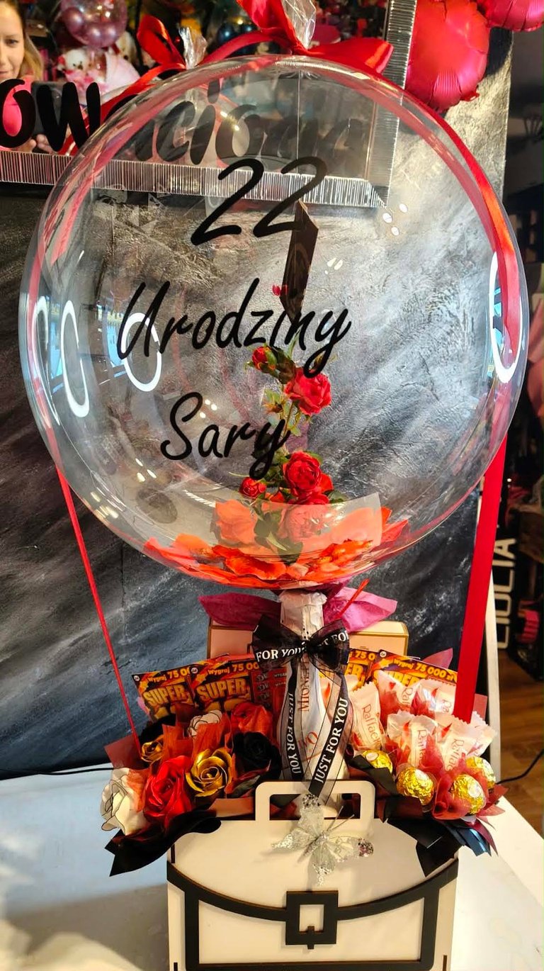 Duży przezroczysty balon-bąbelek z czerwonymi różami w środku, złotym pyłem i napisem Happy 24 Urodziny Saryl, osadzony na ozdobnym pniu z czerwonymi wstążkami, loteriami Super Szans 75 000 zł i czekoladkami Daj Sid