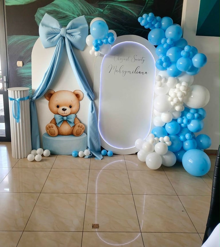 Ścianka baby shower z dużą błękitną kokardą, brązowym misiem w niebieskiej muszce, podświetlaną owalną planszą Chrzest Święty Maksymiliana oraz niebiesko-białym łukiem balonowym