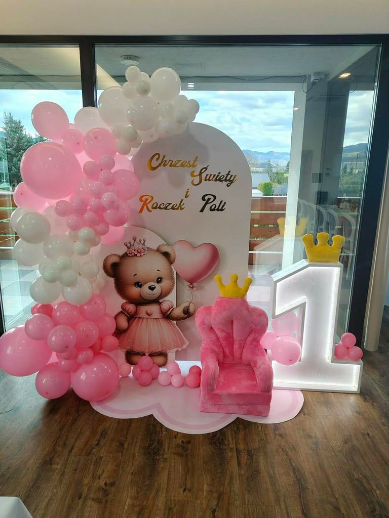 Ścianka Roczek Poli z różowo-białym łukiem balonowym, brązowym misiem w różowej sukience z koroną i sercem, podświetlaną białą cyfrą 1 z żółtą koroną oraz różowym tronem