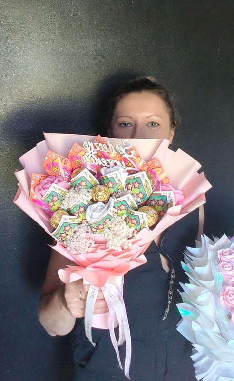 Woman holding a large pink bouquet decorated with colorful candy wrappers, ribbons, and gold accents against a dark background