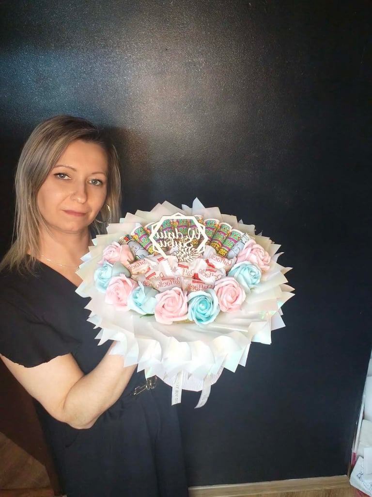 Kobieta w czarnej koszulce trzymająca dekoracyjny bukiet z różowymi i niebieskimi mydlanymi różami oraz kolorowymi batonami, na ciemnym tle