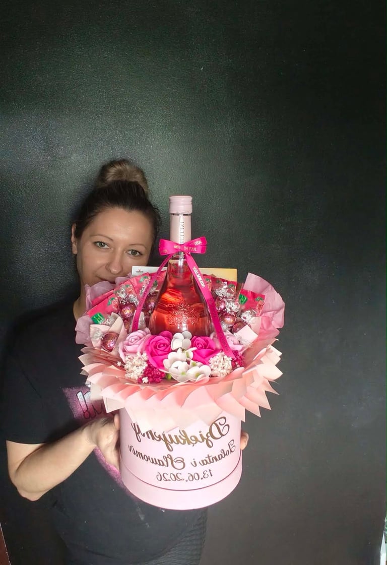 Duży różowy flowerbox z różyczkami, słodyczami, różową wstążką i dedykacją Dziękujemy Romualdowi i Grażynie 13.06.2026, trzymany przez kobietę na czarnym tle