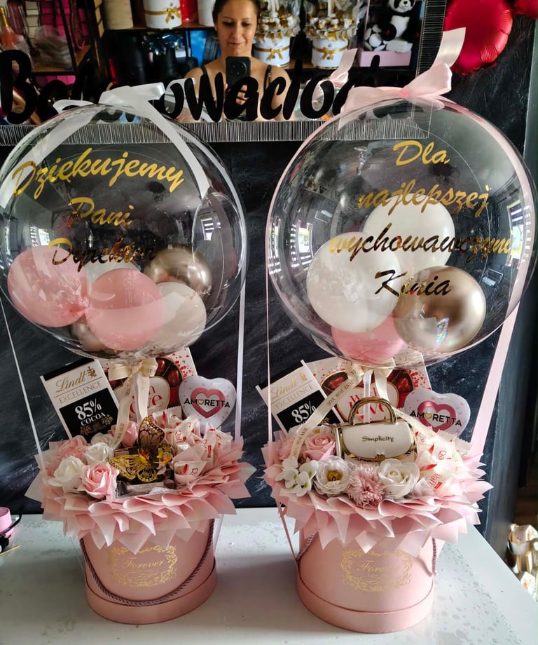 Dwa różowe prezenty z dużymi przezroczystymi balonami-bąbelkami wypełnionymi białymi i złotymi balonami, z dedykacjami Dziękujemy Dla Najlepszej wychowawczyni Pani Doroty i Kinii, czekoladkami Lindt Excellence 85%, Amorettą i torebeczką Simplicity