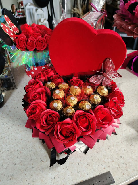 Red heart-shaped arrangement with red roses and gold chocolate candies, red heart balloon on top, displayed on white surface
