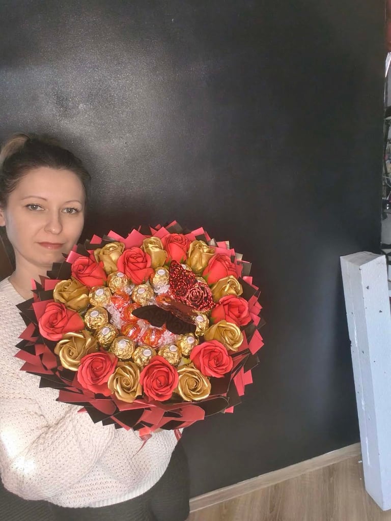 Kobieta trzymająca duży bukiet z czerwonych i złotych mydlanych kwiatów z czekoladkami Raffaello, przewiązany bordową i czerwoną wstążką, na czarnym tle