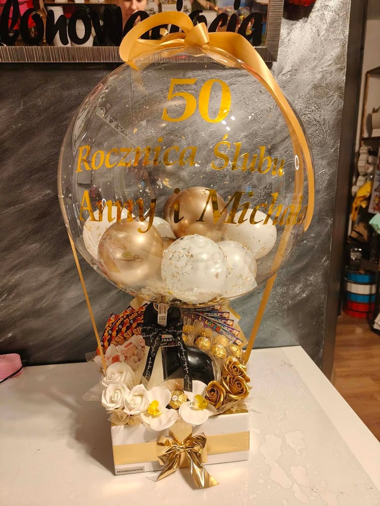 Duży przezroczysty balon-bąbelek z dedykacją 50 Rocznica Ślubu Anny i Michała, wypełniony złotymi i białymi balonami z brokatem, pod spodem czekoladkami Lindt Excellence, Snickers, Raffaello, loteriami i złotymi oraz białymi różami z orchideą