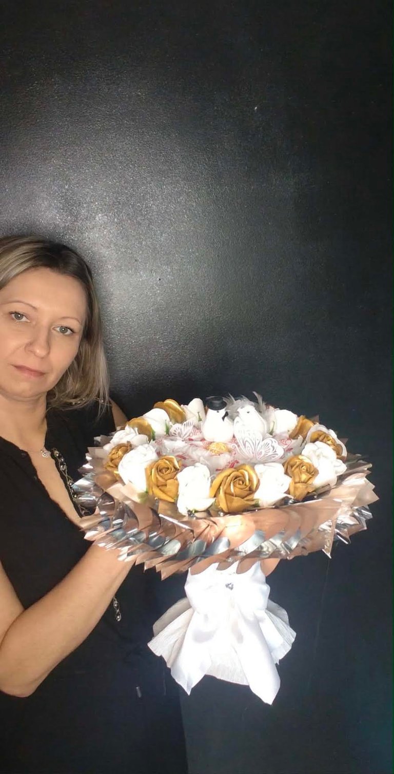Kobieta w czarnej bluzce trzymająca luksusowy bukiet ze złotych i białych mydlanych kwiatów z koronkową różową wstążką u dołu, na ciemnym tle