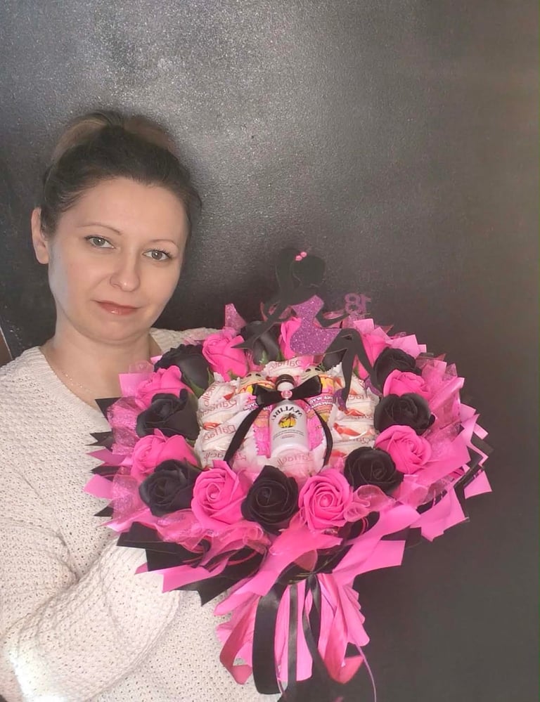 Kobieta w białym swetrze trzymająca duży bukiet z różowych i czarnych mydlanych kwiatów z różowymi wstążkami, kokardą i drobnymi słodyczami w środku