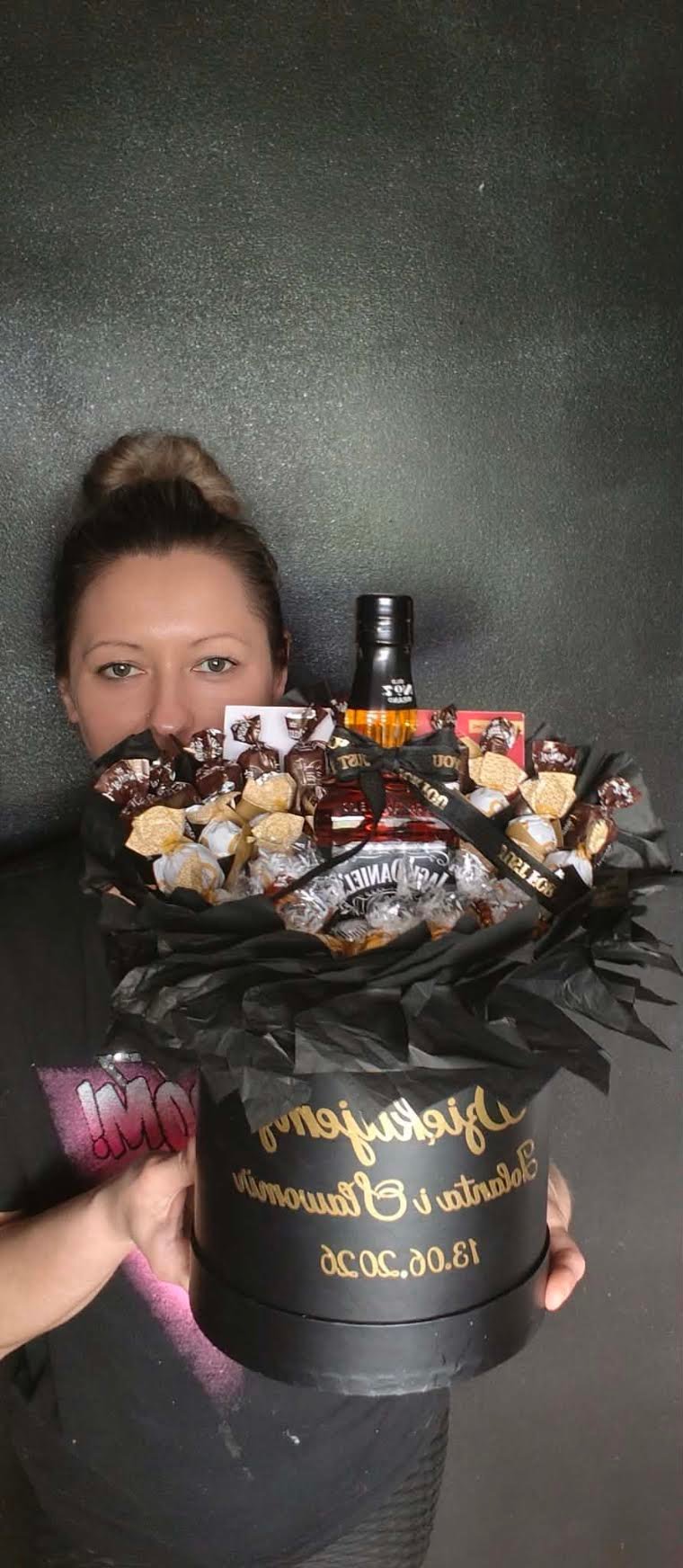 Kobieta trzymająca duży czarny flowerbox z mieszanką czekoladek Zozole, ciasteczek i czarną wstążką z dedykacją Baldwina i Bartka 18.08.20