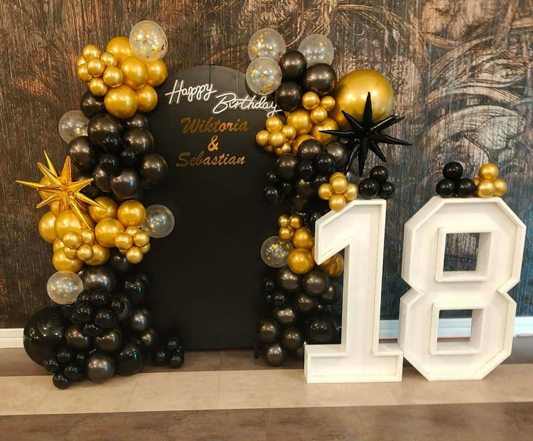 Ścianka 18-stka z czarną planszą Happy Birthday Wiktoria & Sebastian, dużymi białymi cyframi 18 oraz łukiem z czarnych i złotych balonów ze złotą gwiazdą