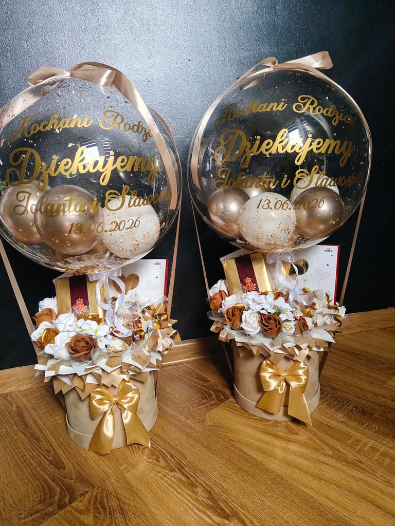 Dwa przezroczyste balony-bąbelki z brązowymi i złotymi perełkami z dedykacjami Kochanej Rodziny Dziękujemy Jolanta i Sławomir 13.06.2026, na złotych cylindrycznych bazach z kremowymi różami, białymi stokrotkami, brązowymi różami i czekoladkami