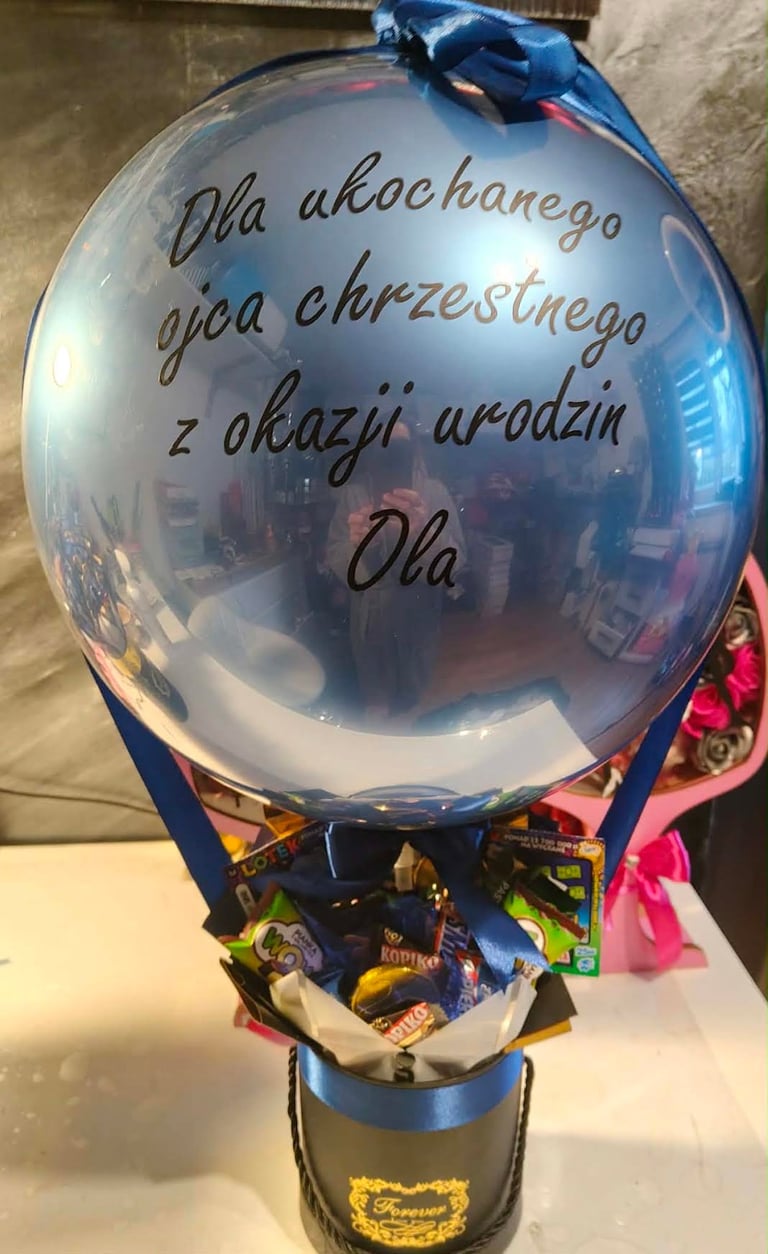 Niebieski duży przezroczysty balon z dedykacją Dla ukochanego ojca chrzestnego z okazji urodzin Ola, z błękitną wstążką u góry, pod spodem slodycze Kopoiko, Kupa i Nosh Bon, w srebrno-niebieskim cylindrycznym pudełku Forever