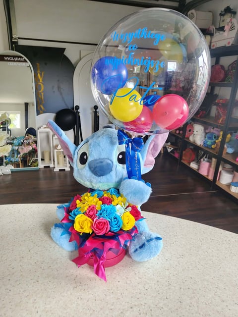 Niebieski pluszowy Stitch trzymający przezroczysty balon-bąbelek z napisem Wszystkiego Najpiękniejszego Pati, wypełniony kolorowymi balonami, z tęczowymi kwiatami u podstawy