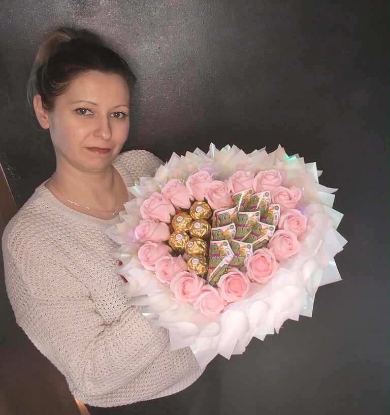 Kobieta w różowym trzymająca serce z różowych mydlanych kwiatów z czekoladkami Raffaello i Daj Sid w środku, w białym opakowaniu z różową wstążką