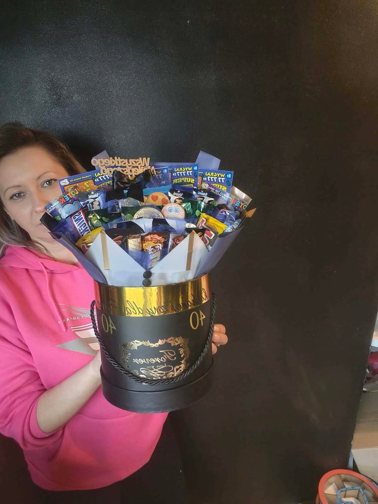 Kobieta w różowej bluzie trzymająca czarno-złoty cylindryczny flowerbox 40 wypełniony loteriami Super Szans i mieszanką słodyczy, z okrągłym okrągłym czarnym napisem Wszystkiego Najlepszego