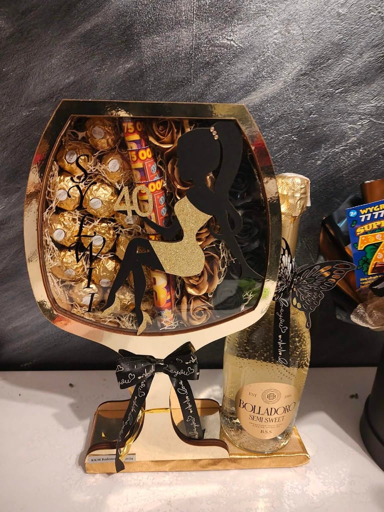 Złoty flowerbox w ozdobnym biało-złotym pudełku z czarną wstążką Love You, wypełniony złotymi Ferrero Rocher, złotymi różami i złotymi loteriami, z czarną kokardą z motylkami