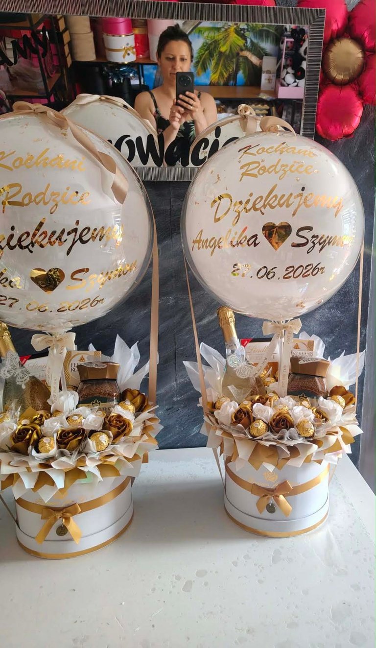 Dwa biało-złote prezenty Kochanym Rodzicom Dziękujemy Angelika i Szymon 27.06.2026r z przezroczystymi balonami-bąbelkami ze złotym pyłem, butelkami Lindt Excellence, złotymi Ferrero Rocher, złotymi i brązowymi piankowymi różami w białym i złotym wypełnieniu