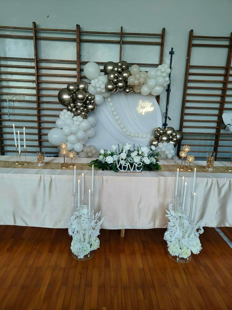 Elegant wedding backdrop with gold and white balloon arch, wooden wall panels, love sign, and table with white flowers and candles