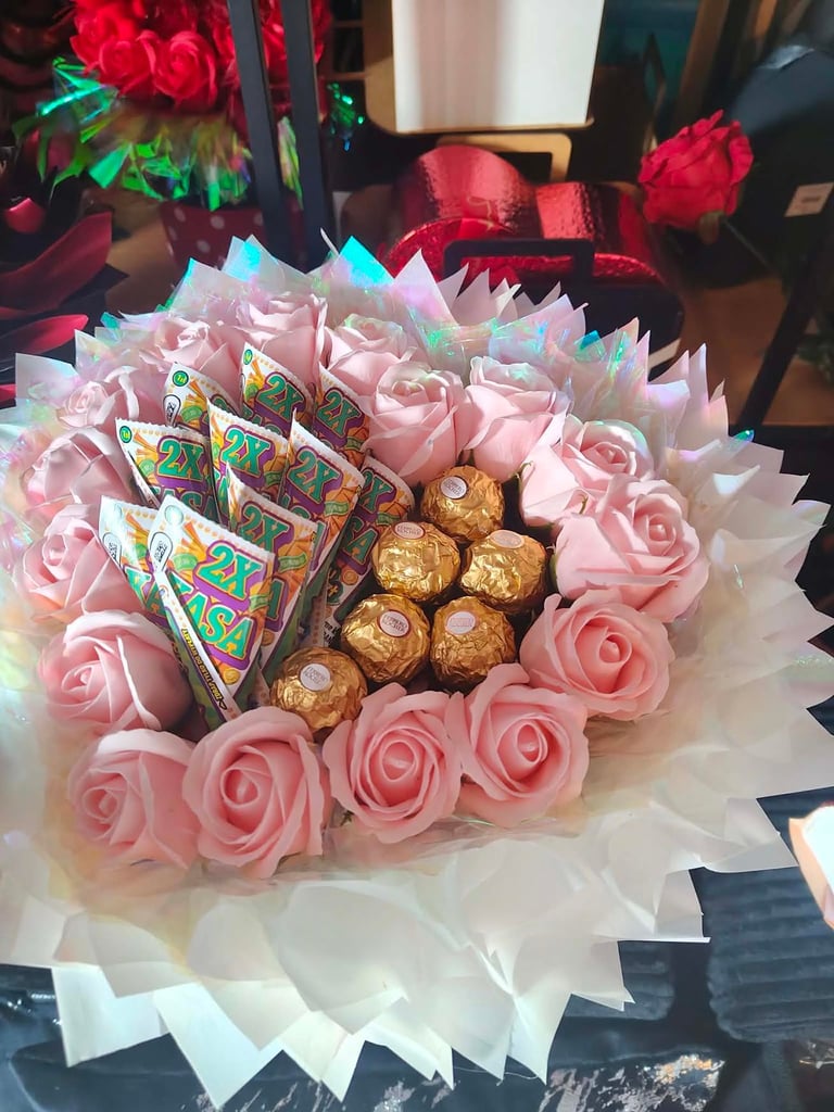 Okrągły bukiet z bladoróżowych mydlanych kwiatów otaczających lody 2X Kasa i złote Ferrero Rocher, w białym opakowaniu z delikatnymi wstążkami