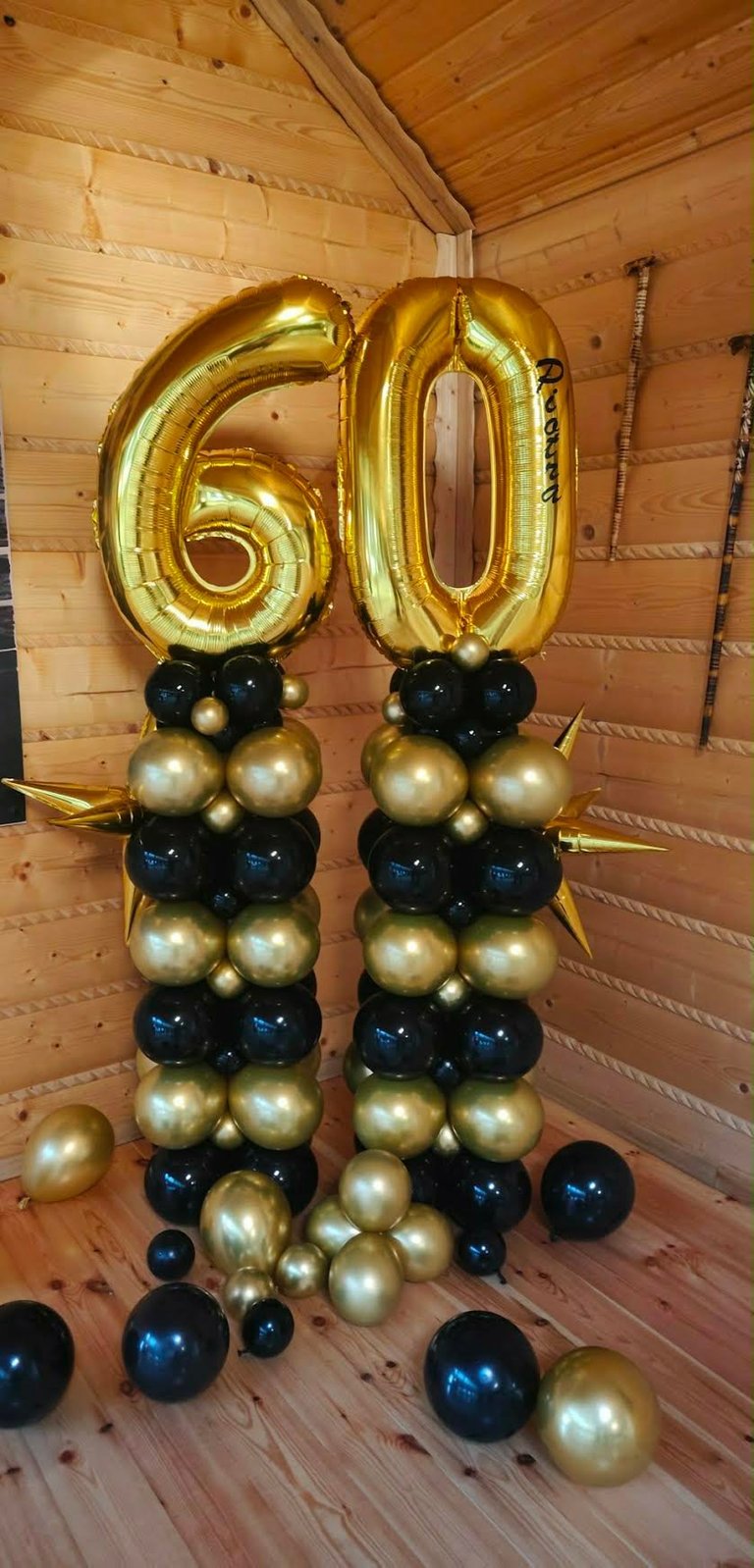 Gold number 60 foil balloons with black and gold sphere balloon stacks against wooden wall for 60th birthday party decoration