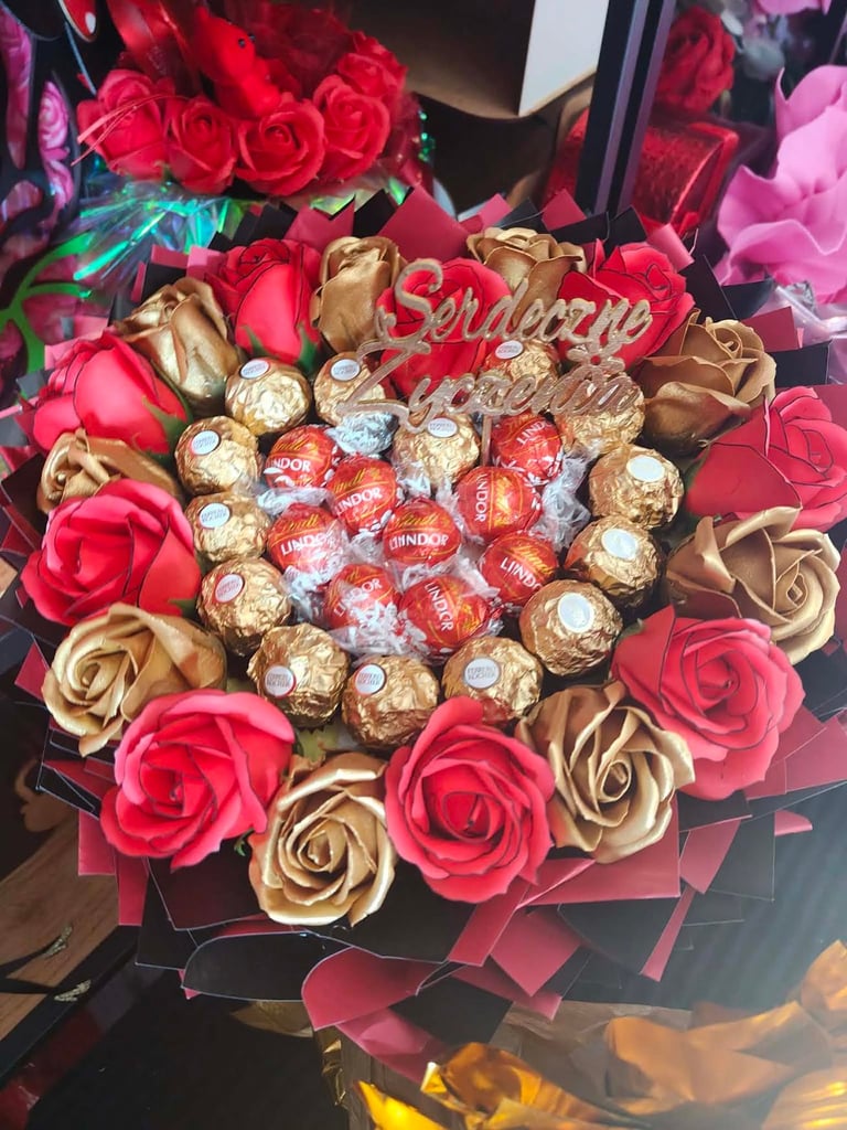 Serce z czerwonych i złotych mydlanych kwiatów otaczających złote Ferrero Rocher, cukierki Lindor i ozdobny złoty napis Serdeczne życzenia pośrodku