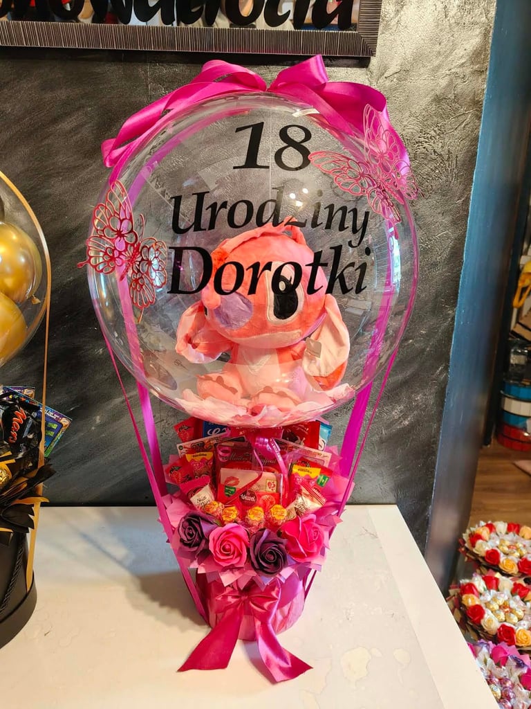 Różowy prezent 18 Urodziny Dorotki z przezroczystym balonem-bąbelkiem z różowymi płatkami róży i różowymi motylkami, pod spodem czerwone i różowe piankowe róże, czekoladkami Wawel, napisem Love You i różową wstęgą