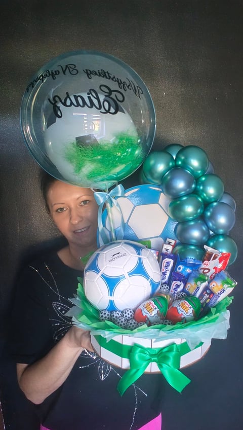 Woman holding a tiered gift arrangement with soccer balls, blue pearl balloons, and a large clear balloon with green tissue paper