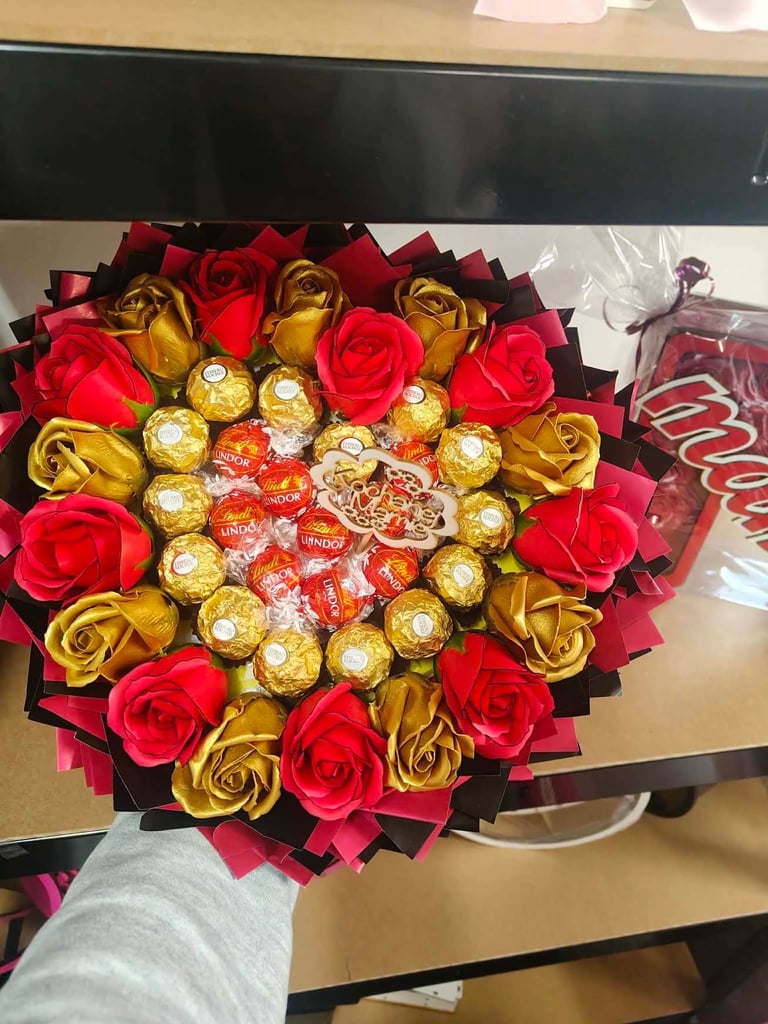 Serce z czerwonych i złotych mydlanych kwiatów otaczających czekoladki Lindor, małe złote pudełeczka i złote serduszko pośrodku, trzymane przez dłoń