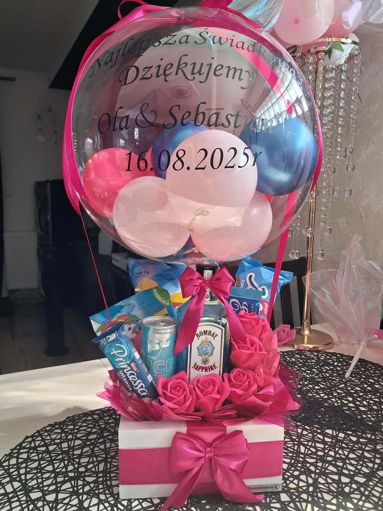 Różowy prezent z dużym przezroczystym balonem-bąbelkiem wypełnionym różowymi balonami, z dedykacją Najpiękniejszego Dnia Dziękujemy Ola & Sebastian 16.08.2025r, z soczkami Tymbark, napojem Havana, różowymi różami i puszką Red Bull