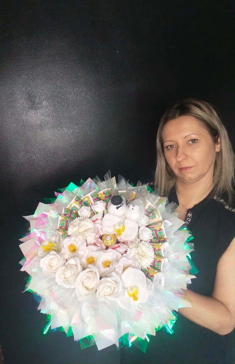 Kobieta trzymająca duży biały bukiet z białych róż i stokrotek z kolorowymi wstążkami, miętowym wypełnieniem i czekoladkami w środku, na czarnym tle