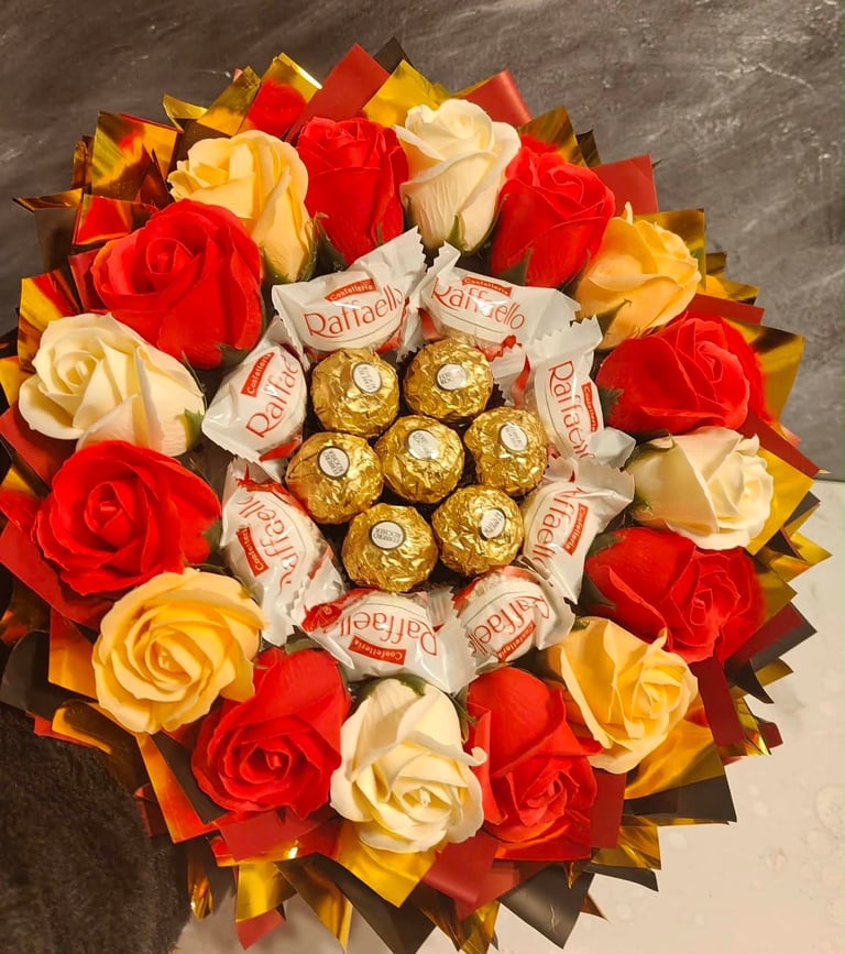 Okrągły bukiet z czerwonych, żółtych i kremowych róż otaczających złote Ferrero Rocher w bordowo-złotym opakowaniu z lustrzanymi wstążkami