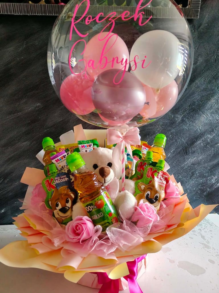 Duży przezroczysty balon-bąbelek z różowymi balonami w środku i napisem Roczek Gabrysi, pod spodem biały miś, soczki HiPP, Kubuś i Kujawianka, otoczone różowym wypełnieniem i różową wstążką