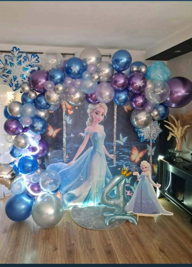 Frozen-themed party decoration with blue, purple, and silver balloons arranged above cardboard cutouts of Elsa and other characters on a wooden floor.