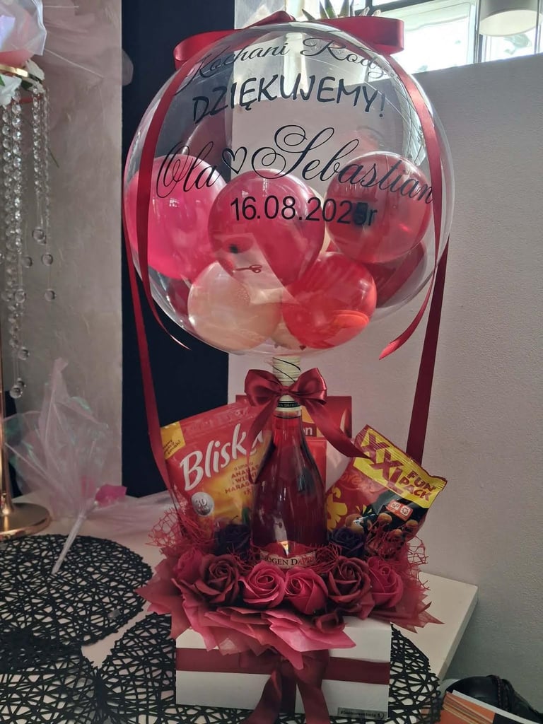 Duży różowy balon z osobistą dedykacją, czerwonymi różami i wstążkami, otoczony czerwonymi pudełkami prezentowymi z pralinami, złotymi dekoracjami i różowym wypełnieniem