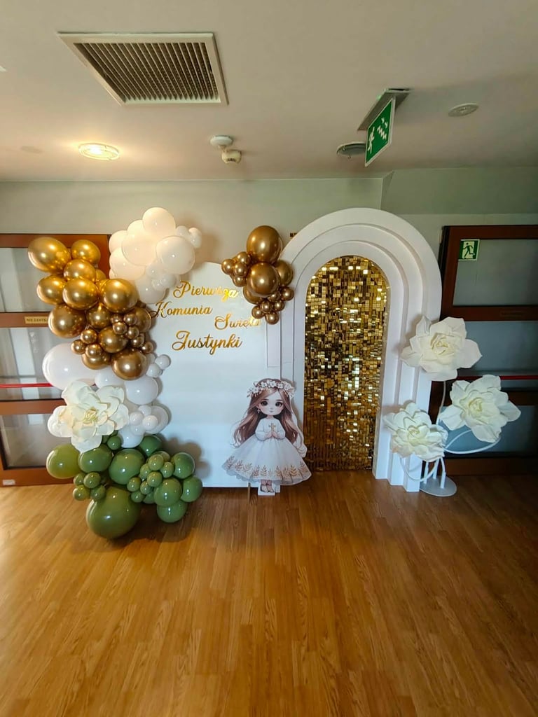 Elegant party decoration setup with white arch, gold and white balloon clusters, white flowers, a doll figure, and green balloons on wooden floor