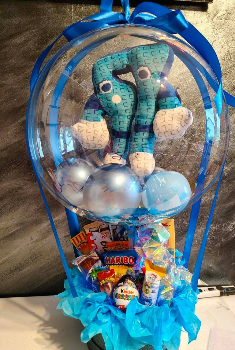 Przezroczysty niebieski balon-bąbelek z pluszowym zielonym dinozaurem w środku, otoczony srebrnymi i niebieskimi balonami oraz słodyczami Dr. Oetker, Haribo, Kinder Bueno i Tic Tac, na błękitnym wypełnieniu z niebieską wstążką