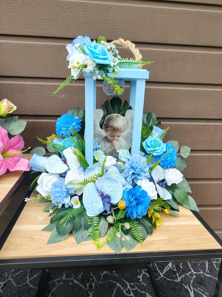 Kompozycja nagrobna z niebiesko-białymi kwiatami wokół jasnoniebieskiej altanki z porcelanową figurką aniołka