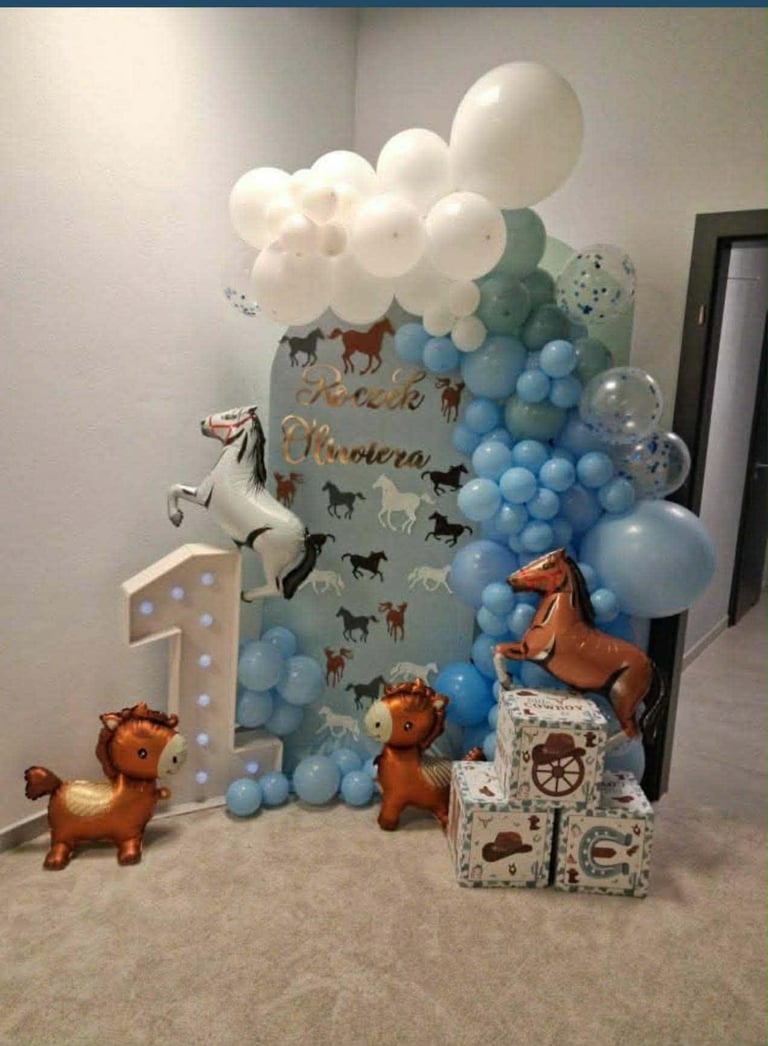 First birthday party backdrop featuring a decorated panel with animal silhouettes, number 1, white cloud balloons, blue balloon garland, and toy horses.
