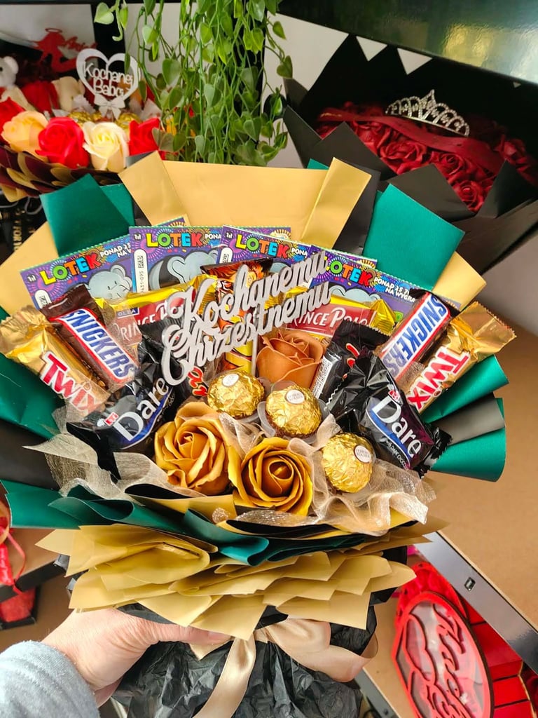 Kolorowy bukiet prezentowy w zielono-złotym papierze z batonami Snickers, Twix, Kinder, wafelkami Prince Polo, loteriami i złotymi Ferrero Rocher, z napisem Kochany Chrześniak, trzymany w dłoniach