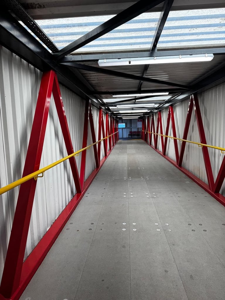 Empty industrial corridor with red metal railings, white walls, skylight roof, and yellow handrail running down the center