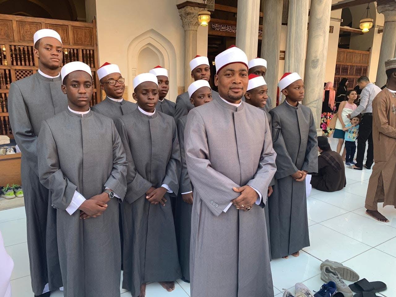 Group of men in gray thobes and white caps standing inside a mosque during prayer