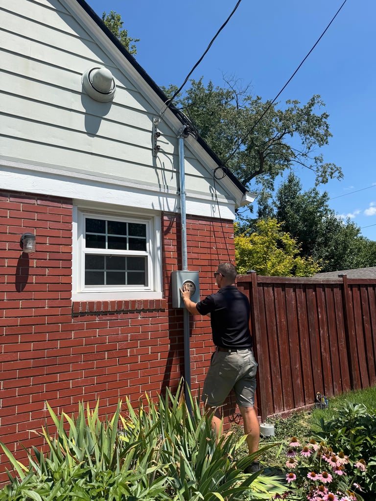 Dylco Electric technician in a black shirt working on an exterior meter/main panel on a brick Central Indiana home — W-2 employees, never subcontractors
